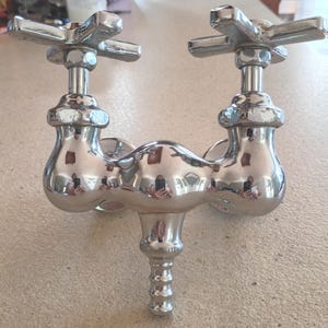 May include: A polished chrome faucet featuring two cross-shaped handles positioned at the top and a central downward-facing spout with a ribbed nozzle. The metal surface is highly reflective, showing distorted reflections of the surrounding environment.