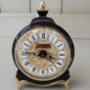 May include: A black and gold Jerger clock with a round face featuring Roman numerals and decorative gold filigree. The clock has a gold base and a gold finial on top. The clock face reads "Jerger" and "West Germany".