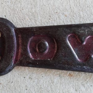 May include: A dark, weathered metal bottle opener featuring the embossed letters D, O, and W across its surface. The opener has a rectangular head with a central cutout and a tapered handle, showing signs of age, oxidation, and dark patina.