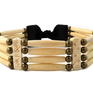 May include: A white bone choker necklace with a black leather tie closure and gold-toned beads. The bone pieces are carved with a geometric pattern.