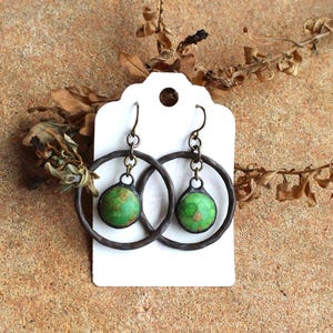 May include: A pair of handcrafted earrings featuring a dark metal hoop design with a small, round, green stone center. The earrings are displayed on a white tag against a textured, tan surface, with dried plant elements.