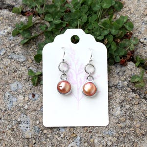 May include: A pair of dangle earrings with peach-colored, round beads. The earrings have silver-colored metal accents and are displayed on a white tag against a textured, gray surface with green foliage.