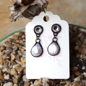 May include: A pair of drop earrings displayed on a white tag. Each earring features a clear, round stone at the top and a pear-shaped, iridescent pearl pendant. The earrings have a dark metal frame.