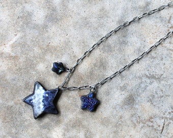 May include: A silver chain necklace with three star-shaped pendants. The largest pendant is white with black marbling, and the two smaller pendants are blue with black accents.