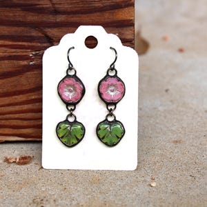 May include: Dangle earrings with a floral and leaf design. The earrings feature pink flower pendants and green leaf pendants, all with black metal detailing. The earrings are displayed on a white tag.