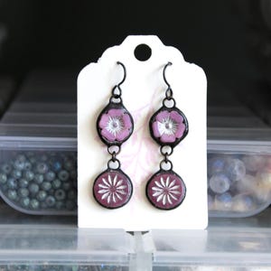 May include: A pair of dangle earrings with a floral design. The earrings feature a dark metal frame with a purple flower and white accents. The lower portion has a circular design with a white starburst pattern. The earrings are displayed on a white tag.