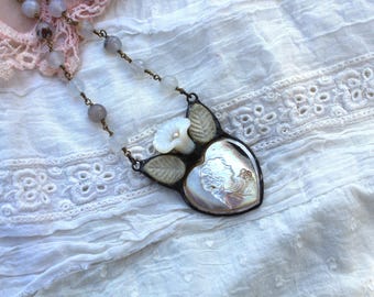 May include: A delicate necklace with a heart-shaped pendant featuring a white flower and a pearl-like shell. The necklace is made of metal and has a vintage aesthetic.