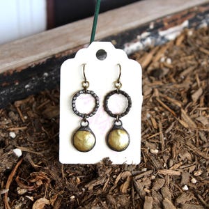 May include: A pair of antique-style earrings. Each earring features a circular design with a textured, dark metal frame and a dangling, gold-colored, teardrop-shaped pendant. The earrings are displayed on a white tag.