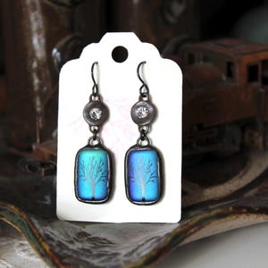 May include: A pair of artisan earrings displayed on a white tag. Each earring features a dark metal setting with a clear crystal accent and a rectangular, blue-green glass pendant with a tree design. The earrings are designed for pierced ears.