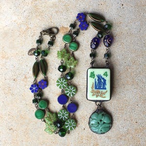 May include: A detailed necklace featuring green, blue, and clear beads. The necklace includes leaf and flower-shaped accents, a rectangular pendant with a blue and white design, and a carved green pendant. The necklace is on a neutral background.
