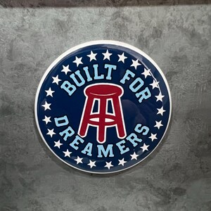 May include: A round, navy blue button with white stars and text. The text reads "BUILT FOR DREAMERS" around a red stool graphic. The button has a white border and is set against a gray background.