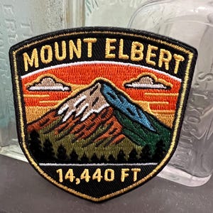 May include: Embroidered patch featuring Mount Elbert, with text reading "MOUNT ELBERT" and "14,440 FT." The patch depicts a mountain scene with a sunset, clouds, and trees. The patch has a black border and a shield shape.