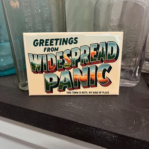 May include: A rectangular magnet with the words "GREETINGS FROM WIDESPREAD PANIC" in a vintage postcard style. The design features a landscape with trees and a sunset. The text "THIS TOWN IS NUTS, MY KIND OF PLACE" is also included.