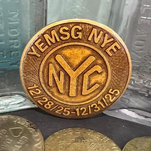 May include: A bronze-colored round token with the text "YEMSG NYE" and "NYC" in a circular design. The dates "12/28/25-12/31/25" are also visible. The token is likely a commemorative item or souvenir.