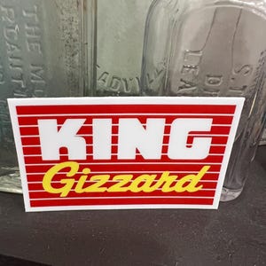 May include: A rectangular sticker with the words "KING GIZZARD" in white and yellow, set against a red background with horizontal white stripes. The sticker is on a dark surface, with blurred glass bottles in the background.