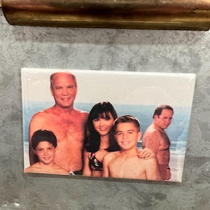 May include: A photo of a family of four on a beach. The father is shirtless and has a beard. The mother is wearing a bikini top and has long dark hair. The two children are both shirtless and have short hair. The photo is printed on a white background and framed in a black frame.
