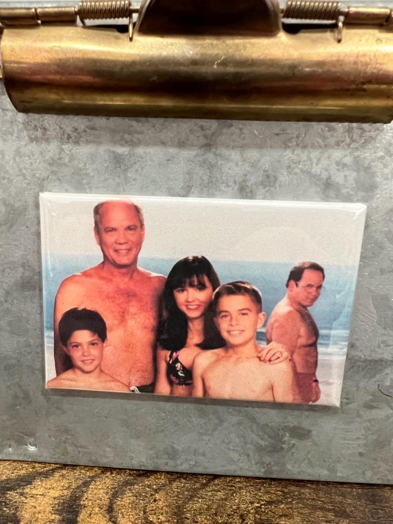 May include: A photo of a family of four on a beach. The father is shirtless and has a beard. The mother is wearing a bikini top and has long dark hair. The two children are both shirtless and have short hair. The photo is printed on a white background and framed in a black frame.
