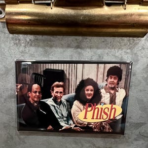 May include: A rectangular magnet featuring a group of four people, with the word "Phish" in a stylized font. The image is set against a backdrop of a room with striped wallpaper. The magnet is attached to a metal clipboard.