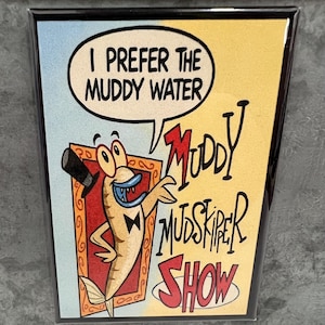 May include: A rectangular magnet with a cartoon image of a fish wearing a top hat and bow tie. The fish is inside a gold frame, with the words "I PREFER THE MUDDY WATER" in a speech bubble. The words "Muddy Mudskipper Show" are also visible.