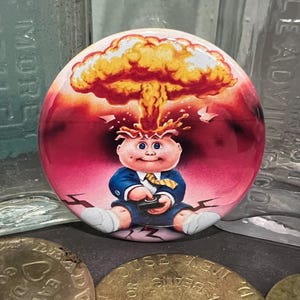 May include: A round button featuring a cartoon baby with a mushroom cloud explosion above its head. The baby is wearing a blue suit and holding a remote. The background is red and the button is surrounded by coins and glass bottles.