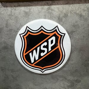 May include: A round, white button with a black and orange shield design. The shield has the letters "WSP" in white. The button has a glossy finish and is likely a pin or badge.