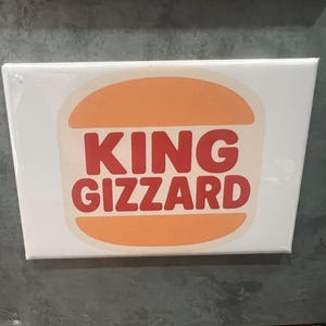 May include: A white rectangular sticker with the words "KING GIZZARD" in red, stylized text. The text is set against an orange, burger-shaped background. The sticker is likely for a band or related merchandise.