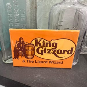 May include: An orange rectangular sign with the words "King Gizzard" in a large, stylized font. Below, the text reads "& The Lizard Wizard." A brown illustration of a figure and barrel is on the left.