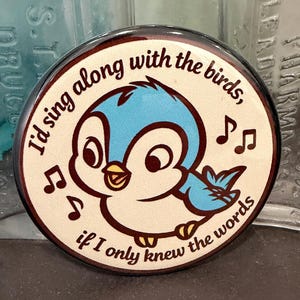 May include: A round, brown-edged button with a cartoon bluebird illustration. The bird is singing, surrounded by musical notes. Text reads, "I'd sing along with the birds, if I only knew the words."