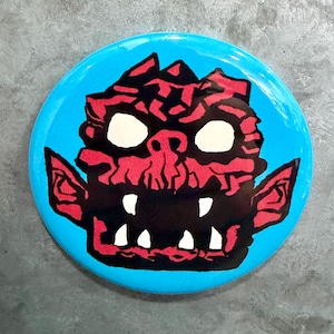 May include: A round, blue button featuring a stylized, cartoonish monster face. The monster has a red face with black accents, large white eyes, and sharp white teeth. The button is a vibrant, eye-catching design.
