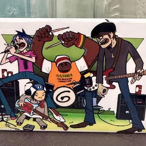 May include: A rectangular magnet featuring a cartoon band performing on a stage. The band members are depicted with exaggerated features, playing instruments and singing. The band's name is not visible.