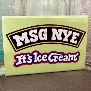 May include: A light green rectangular magnet with the text "MSG NYE" in white letters inside a black and yellow border. Below, the text "It's Ice Cream" is in purple and white. The magnet is displayed against a blurred background.