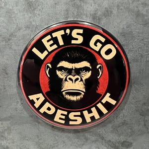 May include: A round button with the text "LET'S GO APESHIT" in white letters on a black background. The button features a stern-faced ape illustration in the center, surrounded by a red circle.