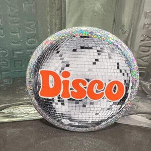 May include: A round, silver disco ball design with the word "Disco" in orange lettering. The border is encrusted with iridescent glitter. The background is blurred, with glass bottles visible.
