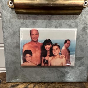 May include: A framed photo of a family of five on a beach. The photo shows a man, a woman, and three children. The man and woman are both wearing swimsuits, and the children are wearing various clothing. The photo is set against a backdrop of blue sky and ocean.