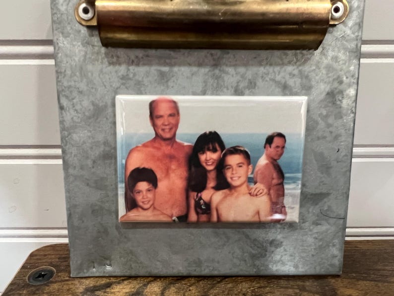 May include: A framed photo of a family of five on a beach. The photo shows a man, a woman, and three children. The man and woman are both wearing swimsuits, and the children are wearing various clothing. The photo is set against a backdrop of blue sky and ocean.