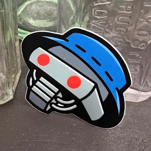 May include: A sticker featuring a robot head design. The robot has a blue head, gray face, and two red circular eyes. The sticker is outlined in black and has a white border.