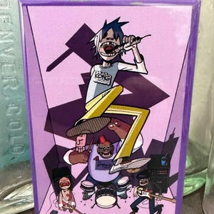 May include: A rectangular purple magnet featuring a cartoon band performing on stage. The lead singer is wearing a white shirt with "Kong Studios" printed on it, yellow pants, and black shoes. The band members are playing drums, bass, and guitar.