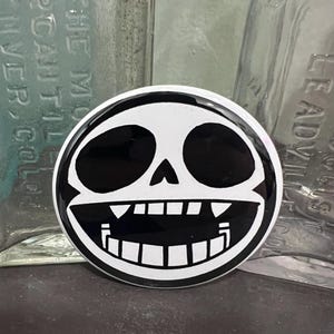 May include: A round, black and white button featuring a cartoon skull design. The skull has large black eye sockets, a wide grin with teeth, and a white outline. The button is likely a pin or badge.