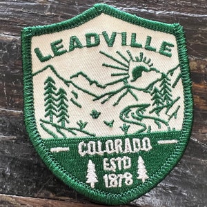 May include: Green and white embroidered patch with the text "Leadville, Colorado, Est. 1878" and a mountain scene with a sun.
