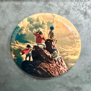 May include: A round button featuring a colorful illustration of a group of cartoon characters on a mountain peak, set against a cloudy sky. The characters are in various poses, with one wearing a red cape.