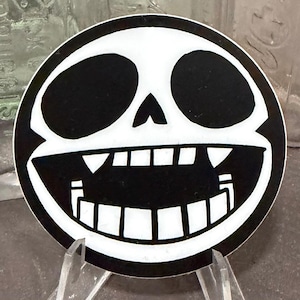May include: A round, black and white sticker featuring a stylized skull design. The skull has large, oval eye sockets, a triangular nose, and a wide, toothy grin. The sticker is displayed on a clear stand.