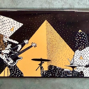 May include: A rectangular magnet featuring a band illustration. Two figures in black and white polka dot outfits play instruments in front of an orange pyramid shape. The background is black with white dots.