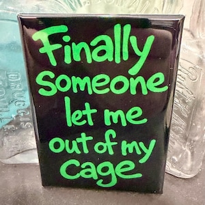 May include: A black rectangular magnet with the words "Finally Someone let me out of my Cage" in bright green, handwritten-style lettering. The magnet is glossy and appears to be on a dark surface.