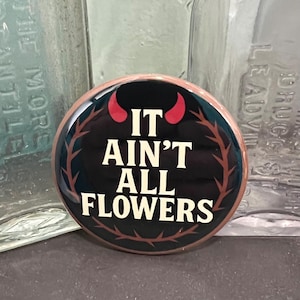 May include: A round button with the text "IT AIN'T ALL FLOWERS" in white letters on a black background. The button has a brown border and a design of red horns and brown branches.