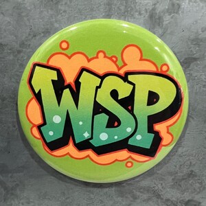 May include: A round, lime green button with the letters "WSP" in a graffiti-style font. The letters are gradient green and black, outlined in black, with orange and red accents. The background features orange cloud-like shapes.