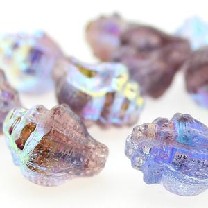 May include: A collection of small, translucent glass beads shaped like seashells. The beads feature a light purple and lavender hue with an iridescent, pearlescent finish that reflects hints of blue and gold light across their textured surfaces.