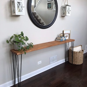 May include: A wooden console table with black hairpin legs. The table is decorated with a potted plant, books, and a framed picture. The table is in front of a white wall with a black round mirror and two white lanterns hanging above it.