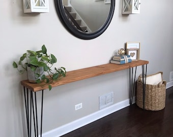May include: A wooden console table with black hairpin legs. The table is decorated with a potted plant, books, and a framed picture. The table is in front of a white wall with a black round mirror and two white lanterns hanging above it.