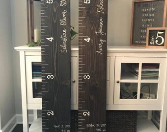 May include: Two wooden growth charts with white painted numbers and names. The chart on the left reads "Sebastian Oliver Strama" and the chart on the right reads "Avery Joanna Kolaczewski". Both charts have the date and time of birth and height measurements in inches.