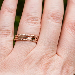 May include: Three gold-toned stacking rings on a finger. The rings have different textures, including a hammered finish and a textured band.